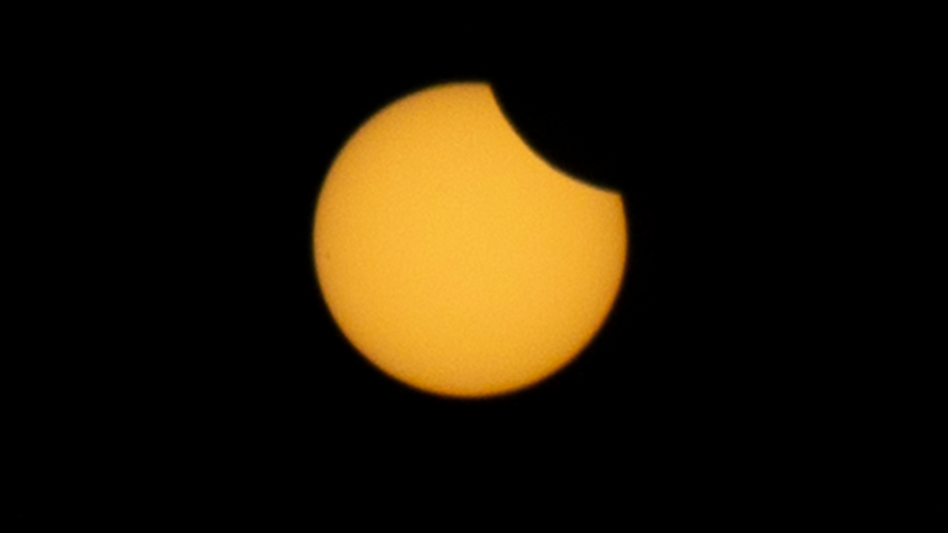 The yellow sun&#039;s disk is shown against a black sky. The curved silhouette of the moon is visible blocking the upper right extreme of its disk.