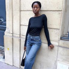 style influencer Sylvie Mus poses on a Paris sidewalk wearing statement earrings, a black boatneck top, high-waisted jeans, and a minimal black shoulder bag