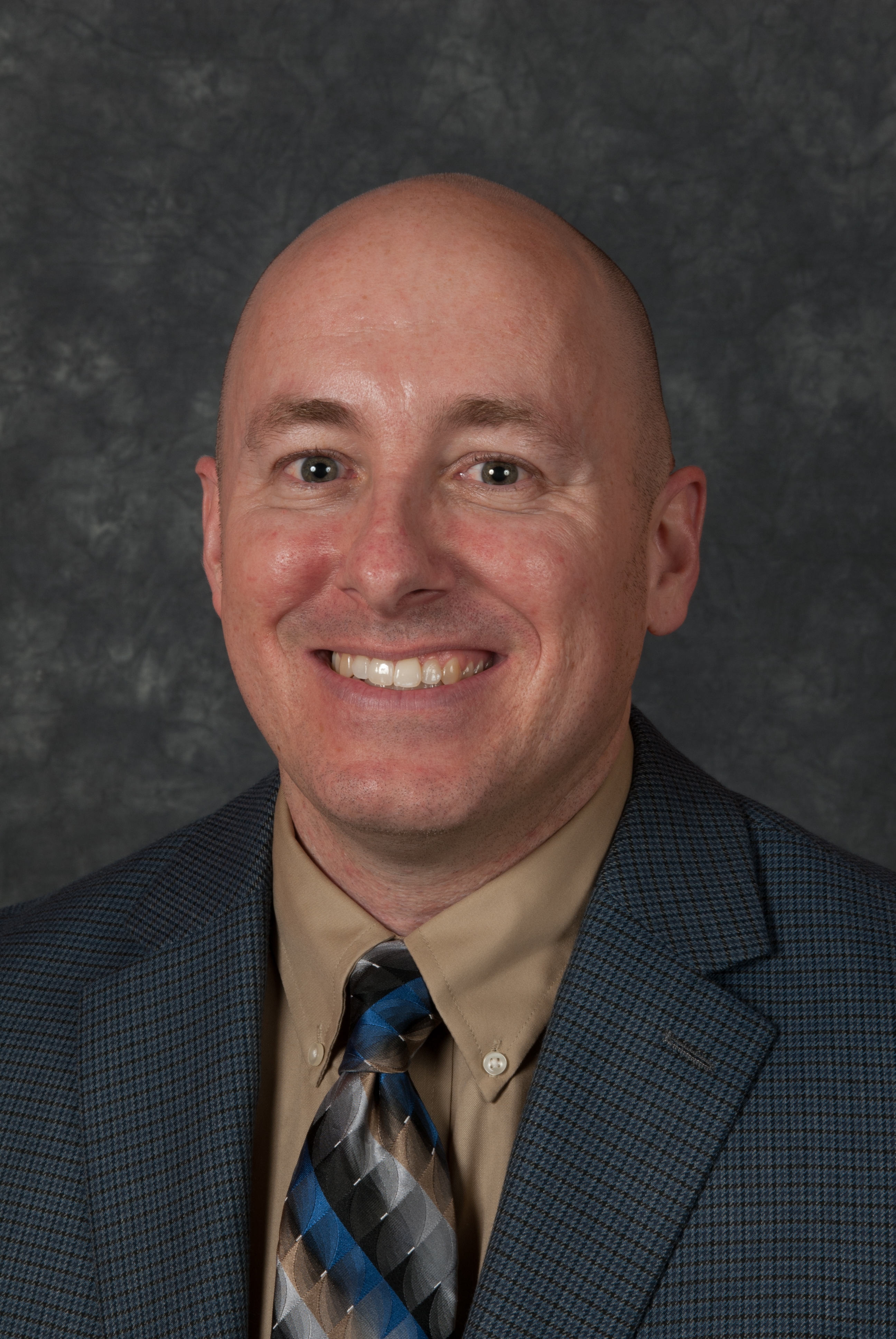 Chad is a white man pictured smiling wearing a dark houndstooth suit, neutral shirt, dark tie, against a mottled dark background in this professional headshot