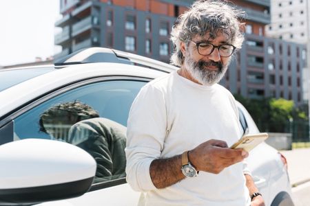 A mature man leans against his car while looking at his phone. 