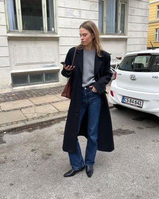 Scandi influencer Clara Dyrhauge poses on the streets of Copenhagen while looking at her phone wearing a long black coat, light gray crewneck sweater, camel bag, dark-wash jeans, and black pointed-toe boots