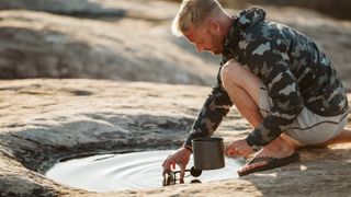 Man in camo jacket collects water to cook with from shallow puddle