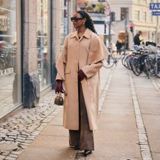 Woman wears trench coat and trousers while posing with a small Louis Vuitton handbag. 