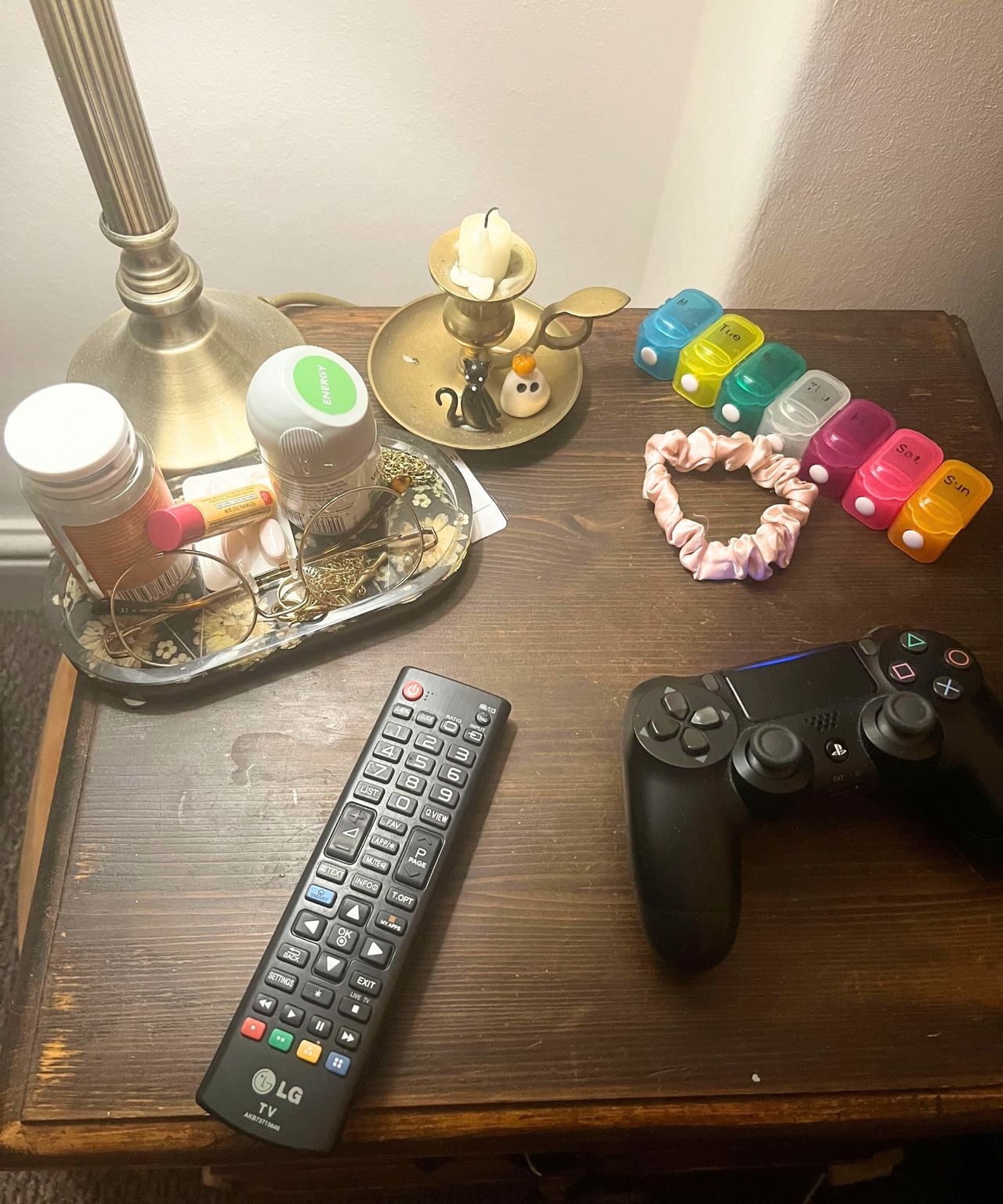 The top of a dark wooden nightstand with a lamp, a pair of glasses, a pill box, and a candle holder, cluttered with a remote, medications, and a hair tie.