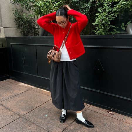 a woman wearing a red cardigan, white tee, black culottes shorts, and black loafers