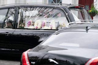 A hearse with a white coffin inside with flowers spelling out 'Lola' in the window.