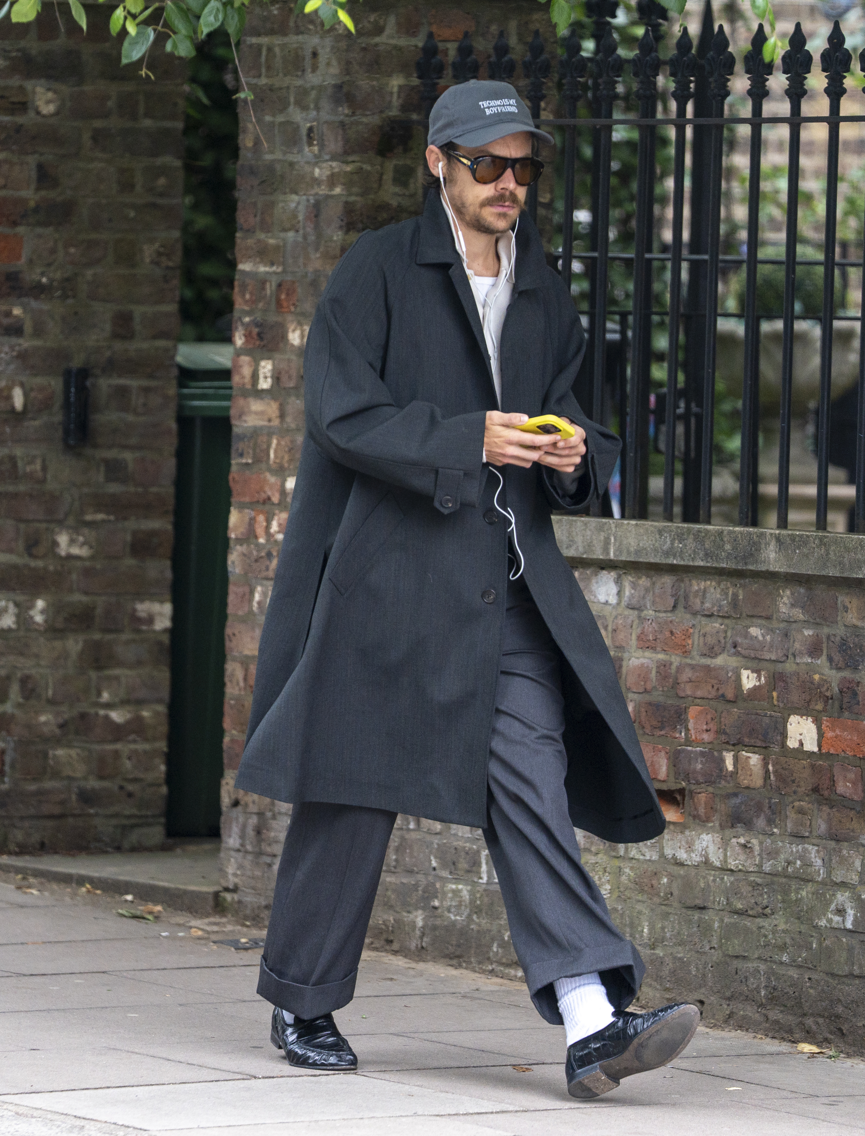 Harry Styles wears black leather loafers with white socks, black straight-leg trousers, a black overcoat and blue button down shirt as he walks through the streets of London.