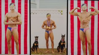 Zac Efron competing in bodybuilding in scene from Baywatch 2017. He's wearing red, white and blue flag suit. 