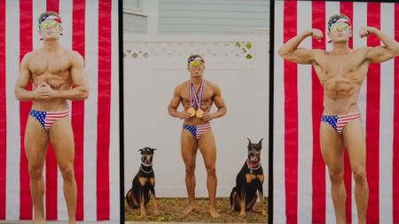 Zac Efron competing in bodybuilding in scene from Baywatch 2017. He's wearing red, white and blue flag suit. 