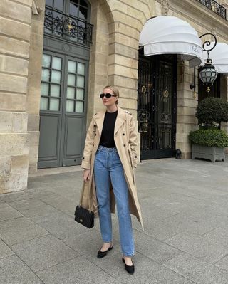 editor mode Kristen Nichols berpose dalam pakaian penuh gaya di jalanan Paris mengenakan kacamata hitam oval besar, jas hujan, atasan hitam dasar, celana jins lurus, tas Chanel, dan sepatu datar slingback suede hitam