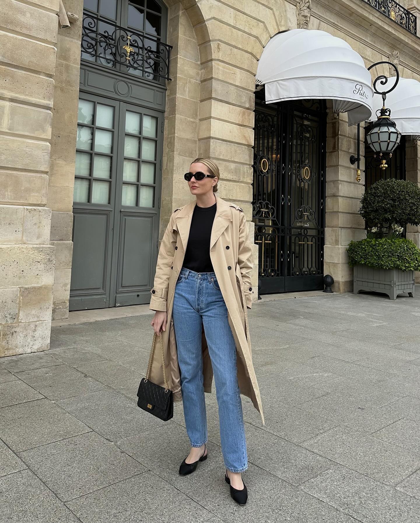 fashion editor Kristen Nichols poses in a stylish outfit on the streets of Paris wearing black oversize oval sunglasses, a trench coat, basic black top, straight-leg jeans, Chanel bag, and black suede slingback flats