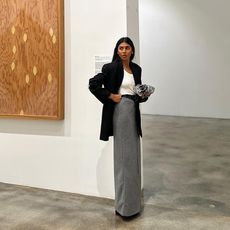 Fashion influencer Monikh Dale in an art gallery wearing a long grey skirt, basic white top, and long black blazer, carrying a clutch handbag.