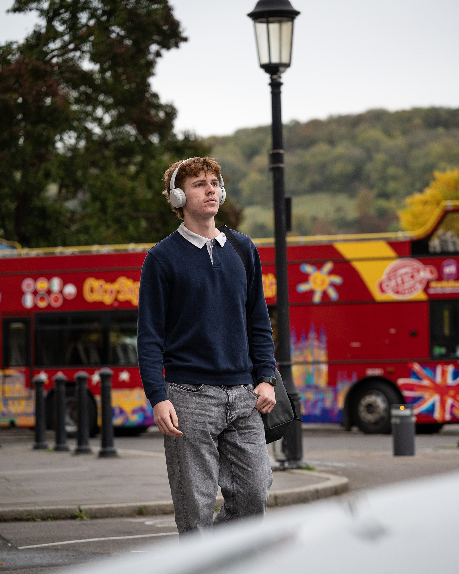 Street photograph of man with headphones next to street lamp with red bus in background