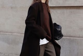 @modedamour wearing a dark, long coat, a burgundy sweater, brown pants, and a black bowler bag.