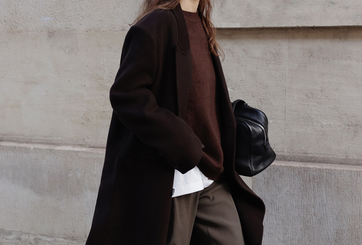 @modedamour wearing a dark, long coat, a burgundy sweater, brown pants, and a black bowler bag.