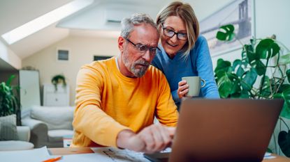 A retired man looks at his laptop with his smiling wife looks over his shoulder.