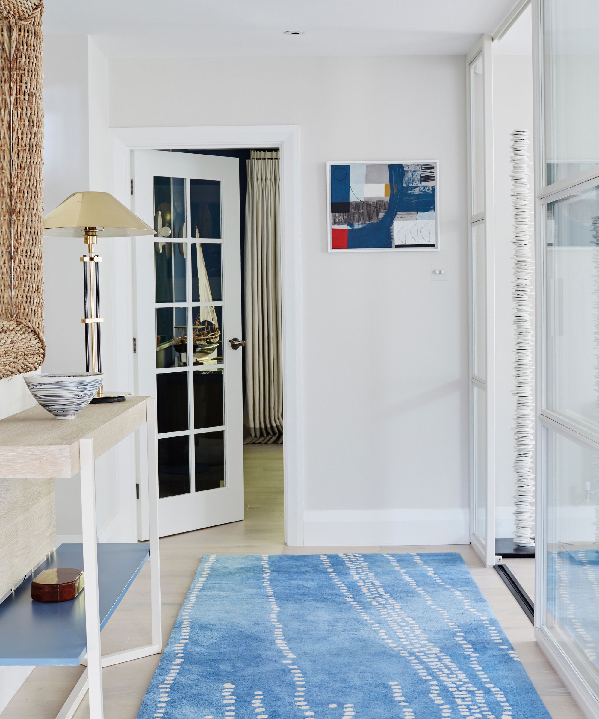 A white entryway with a blue and white rug, side table with a bowl and lamp, and open door with room beyond seen.