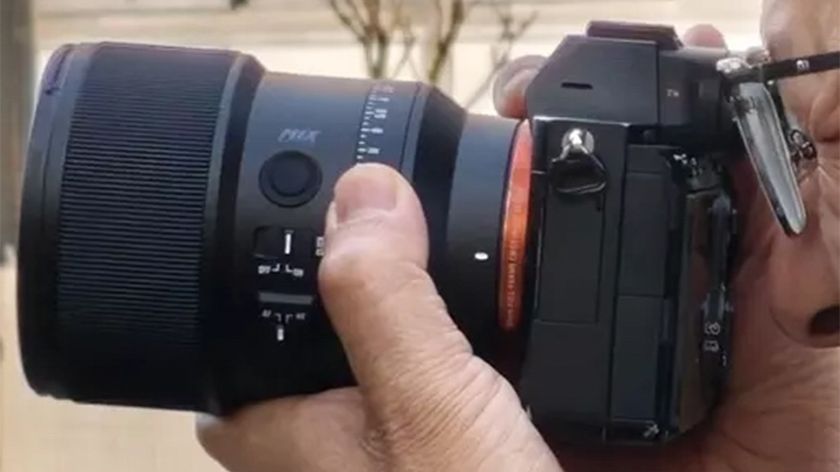 A man wearing glasses takes a picture with a black camera with a black lens attached. 