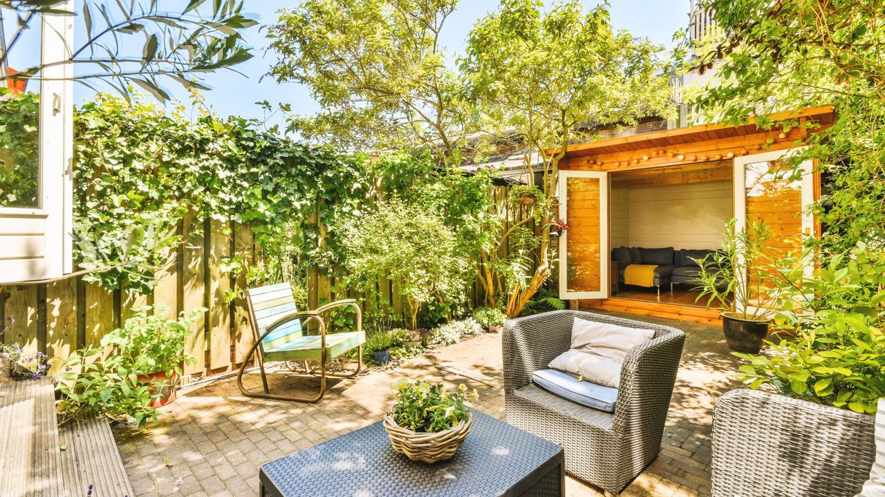 Learning how to repel ticks in your backyard is useful. Here is a backyard space with wooden fencing with vines and trees next to it, two outdoor seats with cushioning, a black rattan coffee table with a planter on it