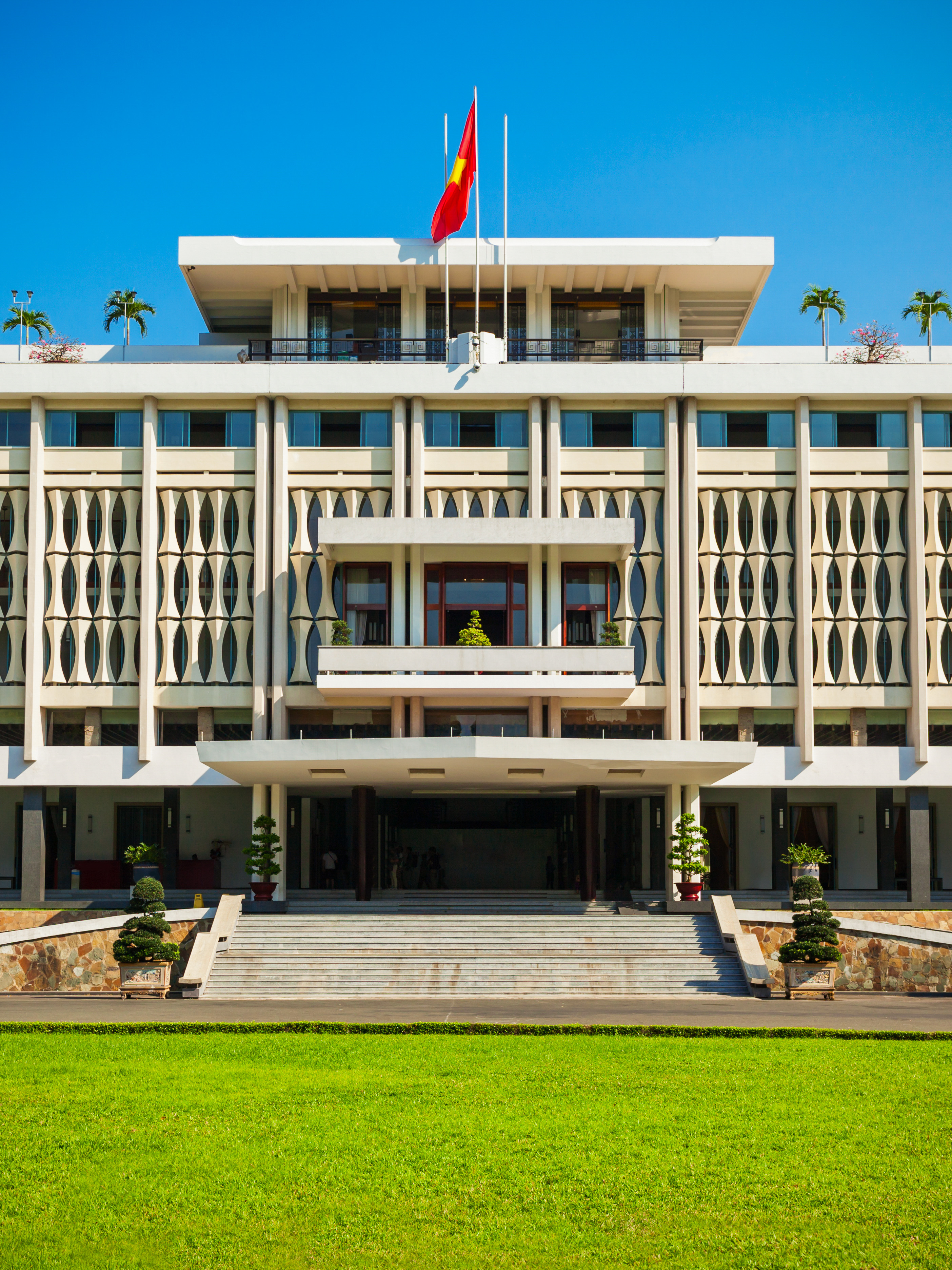 A sunny view of a historical, modernist building with a geometrical facade, a red and yellow flag hanging from its top story, and lush greenery all around it.