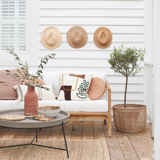 budget garden ideas, porch idea with white wooden walls, neutral scheme with wood and white sofa, concrete and metal coffee table, basket with plant, graphic cushions, straw hats on wall, shutters, jute rug