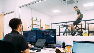 A man in a room on a static bike is watched by another man at a bank of computer screens