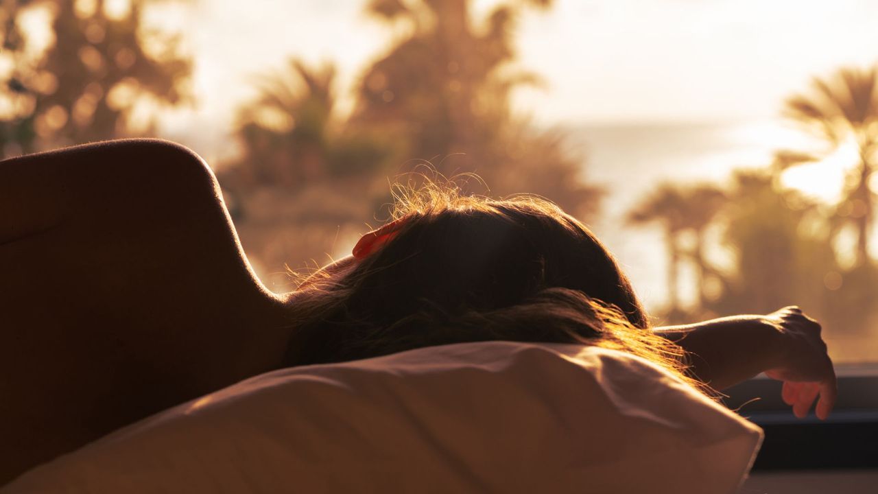 Woman sleeping facing rising sunshine through window with palm trees, representing how to nap in the summer months