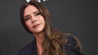 Victoria Beckham smiling with curled long brunette hair and wearing a black blazer