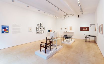 A gallery room with white walls and beige tiled flooring. Multiple framed images on the white wall. In the center of the room are furniture items placed on individual square podiums. A brown chair, A white table with brown square blocks placed on it. A chair with metal frame and black textile and a small glass table with metal base. 