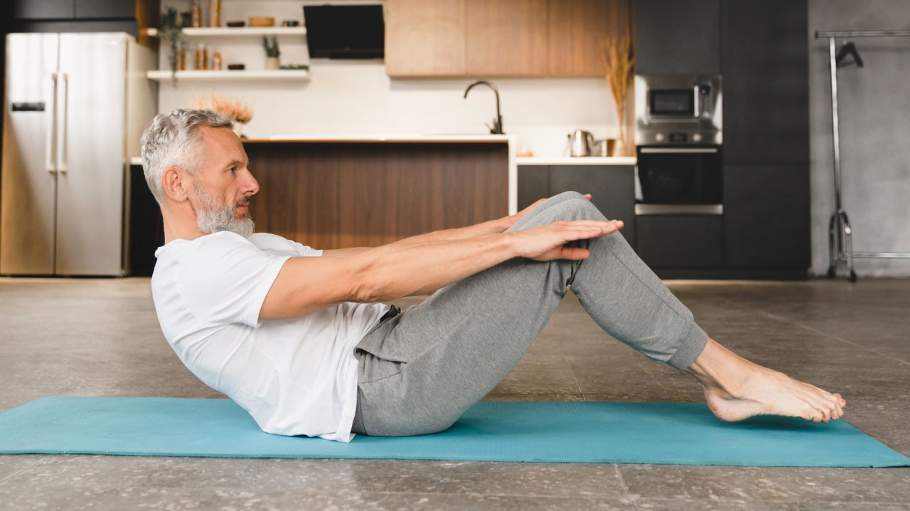 A man performs a variation of the Pilates Hundred on a mat in a kitchen. He is sat on the mat, with his knees bent and feet hovering off the floor. His torso is at a 45° to the mat and his arms are straight out, parallel with the ground. Behind him we see an oven, a fridge and kitchen units. 