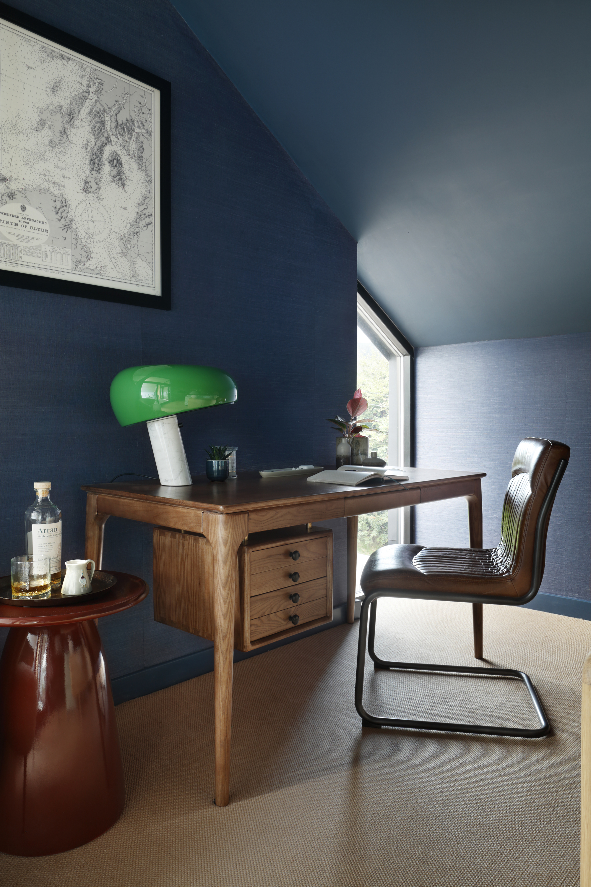 Image of a home office painted dark blue with slanted ceilings. There is a wooden desk with a green and white mushroom lamp on it and a red side table with drinks.
