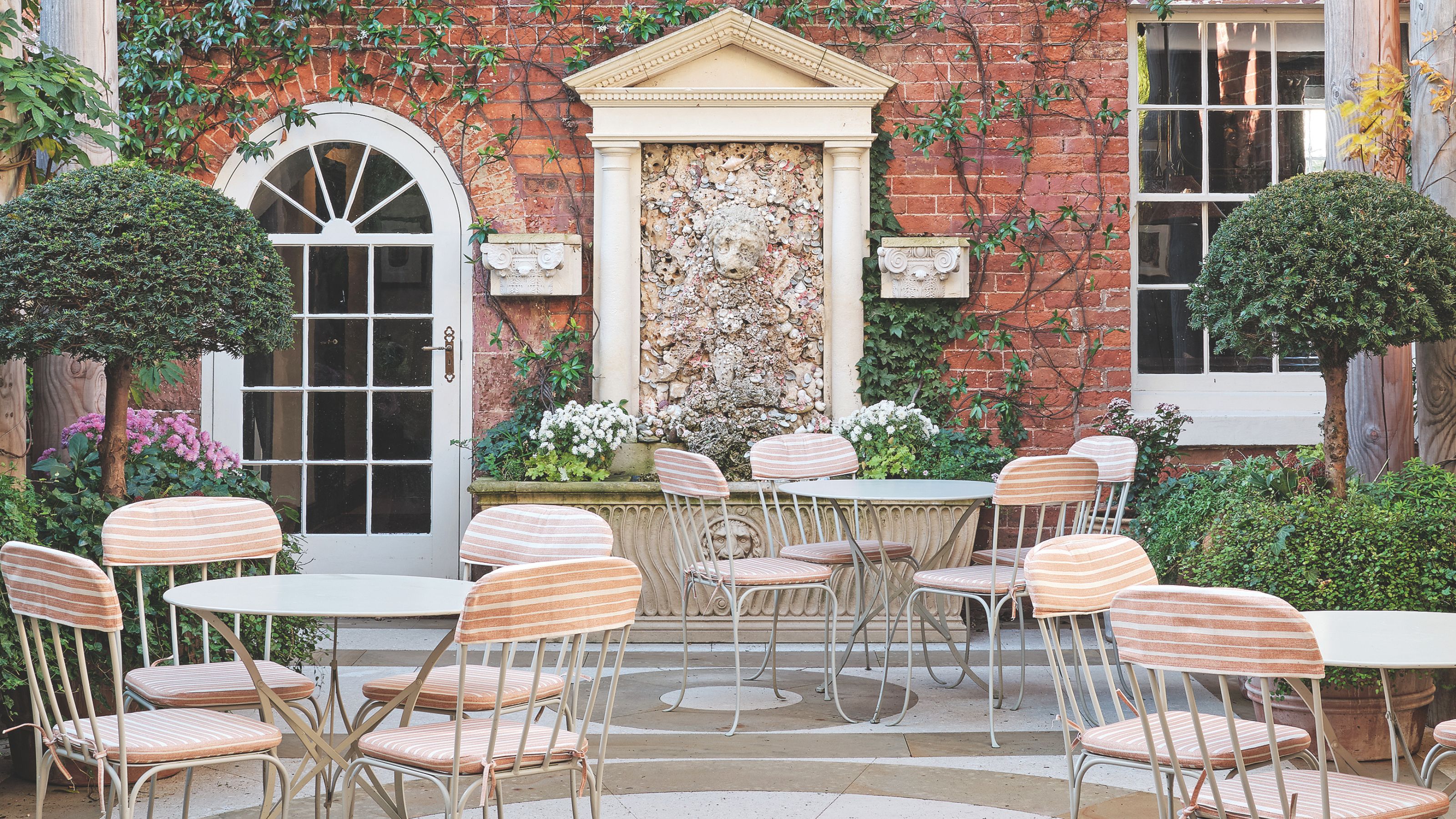 An outdoor brick courtyard featuring a classical stone wall fountain decorated with shells, surrounded by manicured greenery and bistro-style tables with striped cushions