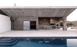 A summer retreat in Greece. A closer look at the outdoor area with a pool and a patio with gray metal seating and a grill, built into the stone wall.