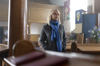 Calder (Ashley Jensen) stands inside a church, still wearing her outdoor coat and scarf. She is talking to someone off-camera.