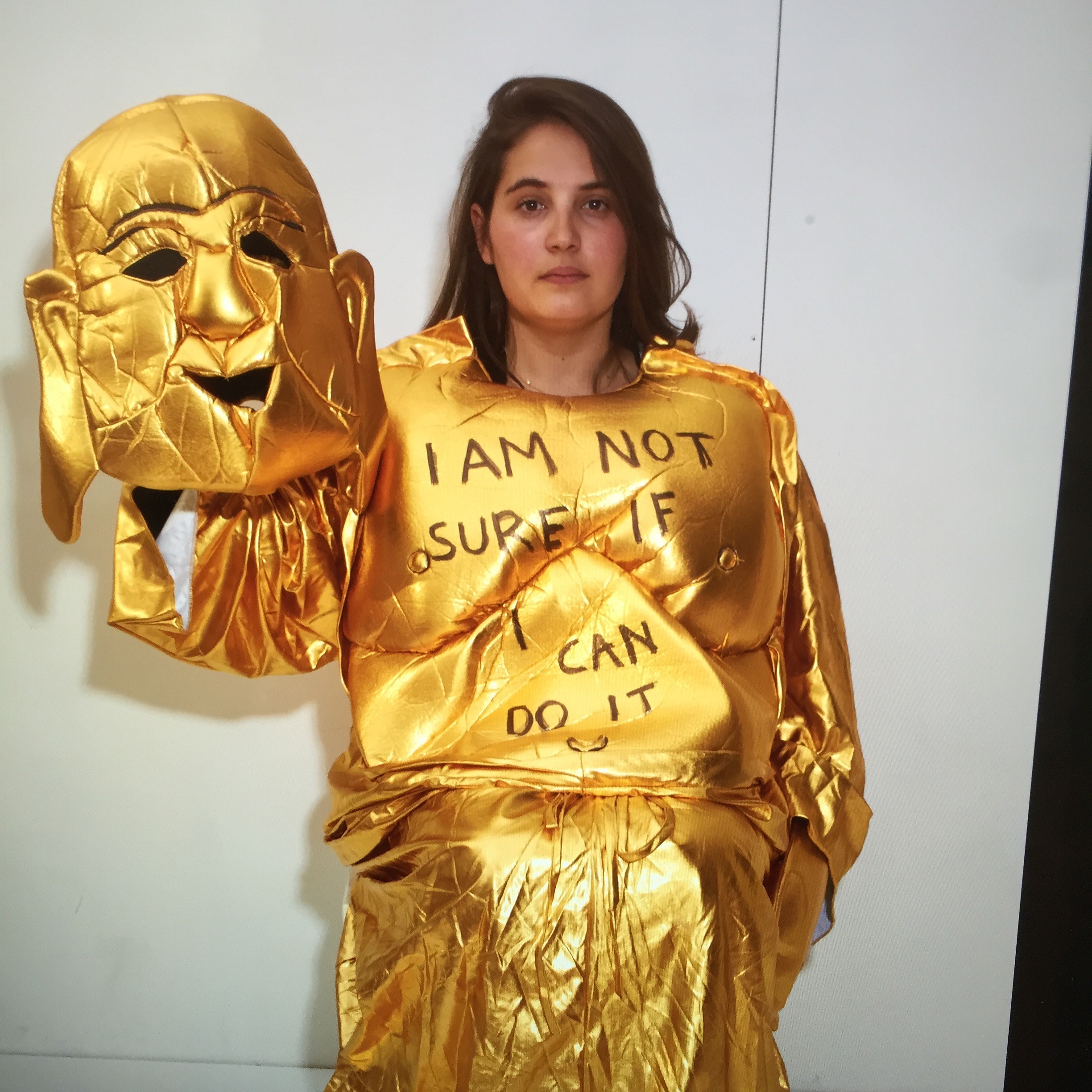 A woman dressed in a golden costume wears a top bearing the message &quot;I am not sure if I can do it&quot; in black while standing against a white wall.