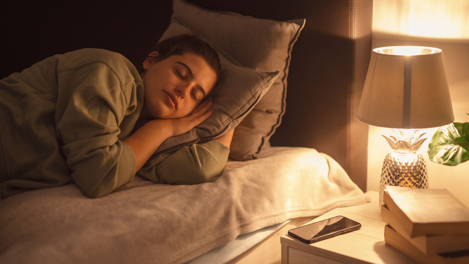 A woman going to sleep in bed at night. She is lying on her side and her bedside lamp is still on.