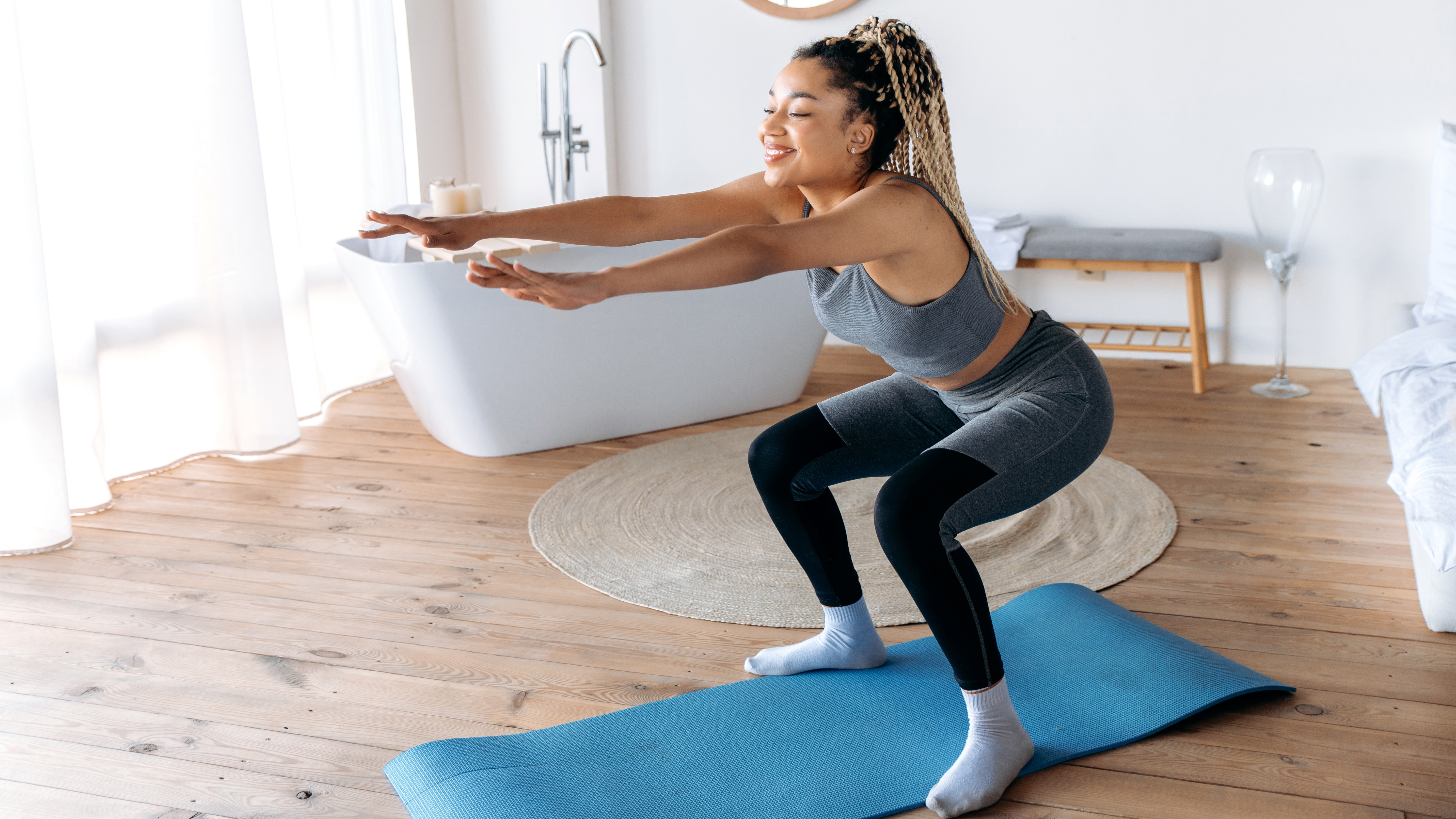 A woman performs a bodyweight squat in a large bathroom on an exercise mat. She is wearing leggings, a sports bra and socks. Behind her we see a free-standing bath, a round mirror and long curtains.