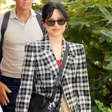 Jenna Ortega wears burgundy loafers, white socks, a checkered blazer, khaki shorts, and a black crossbody bag.