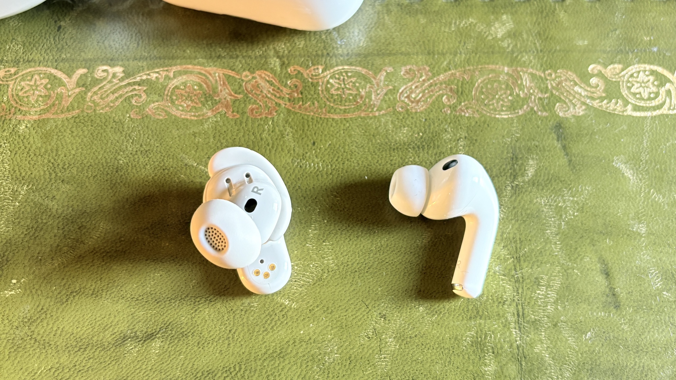 One earbud each of the AirPods Pro 3 and Bose QuietComfort Ultra Earbuds (2nd Gen) on a green surface.