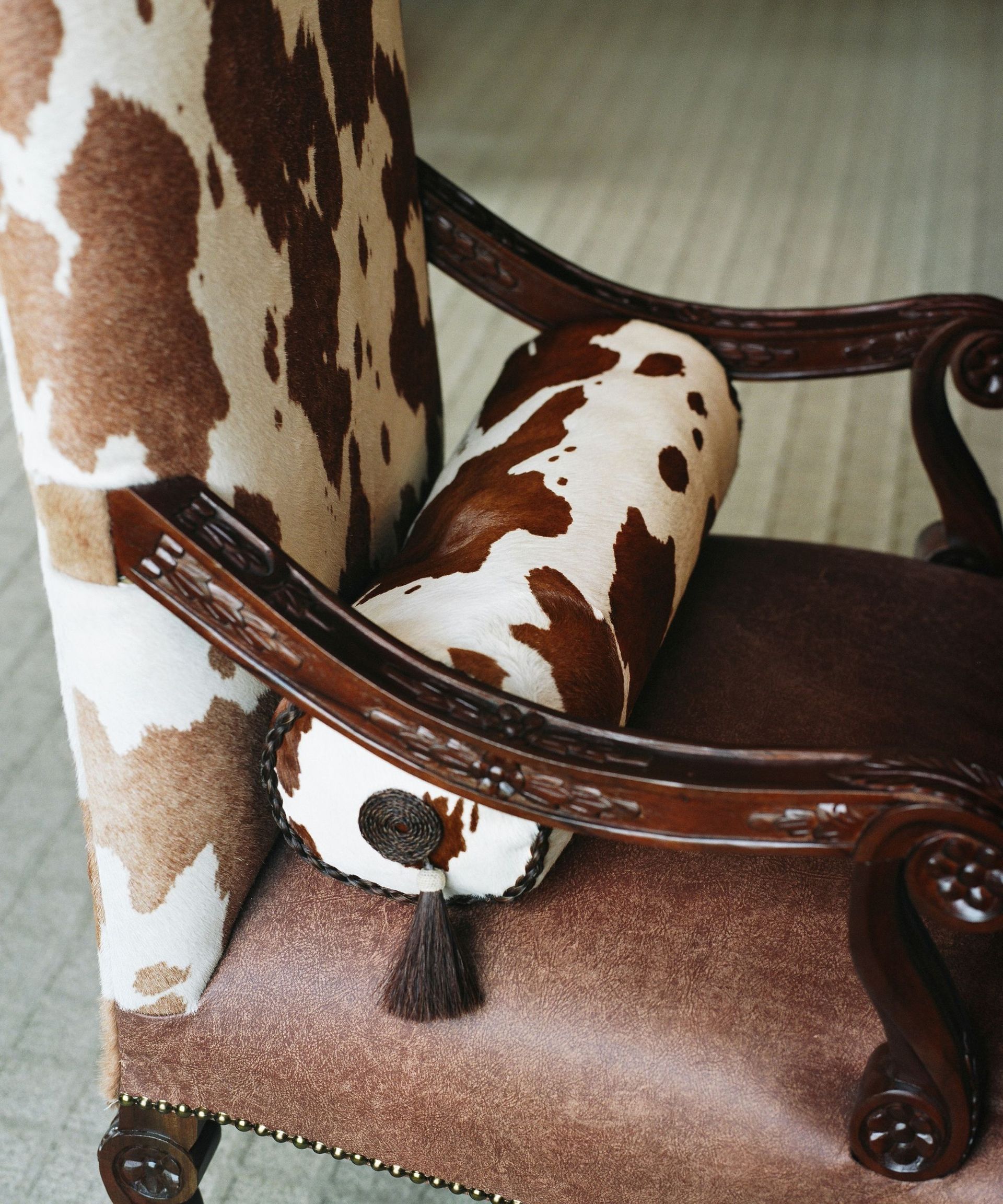 A dining chair upholstered in brown leather and cow print, with a cow print bolster cushion on it