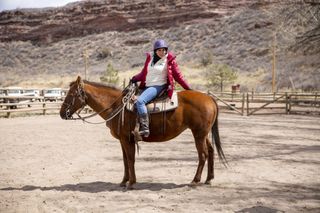 Ruby Wax is sitting on a horse, in a sandy yard in Colorado. She is riding astride the horse and turning to face the side, with one hand resting on the horse's hind.