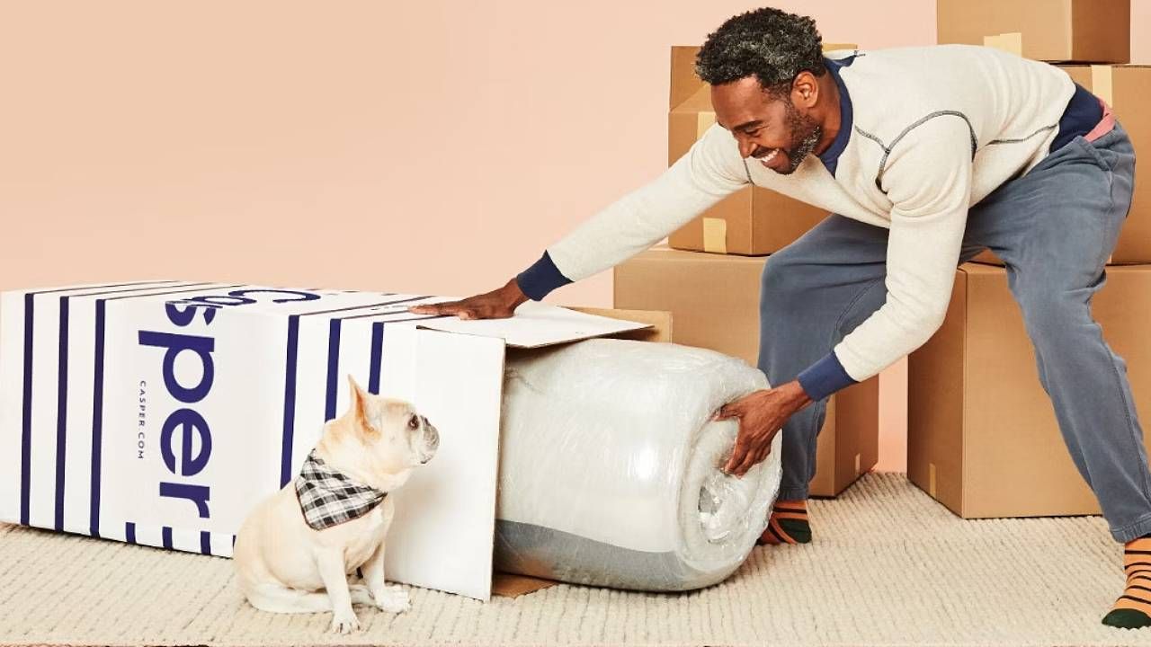 A man pulling a bed in a box mattress out of a Casper box with a dog sat next to it