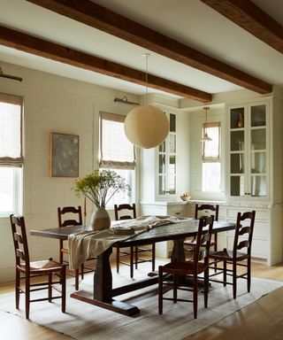 cream beamed dining area with glazed cabinets, wooden trestle table, ladderback chairs and round cream pendant