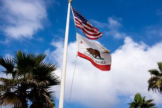 United States flag and California state flag pictured on a flag pole with blue skies and clouds in the background