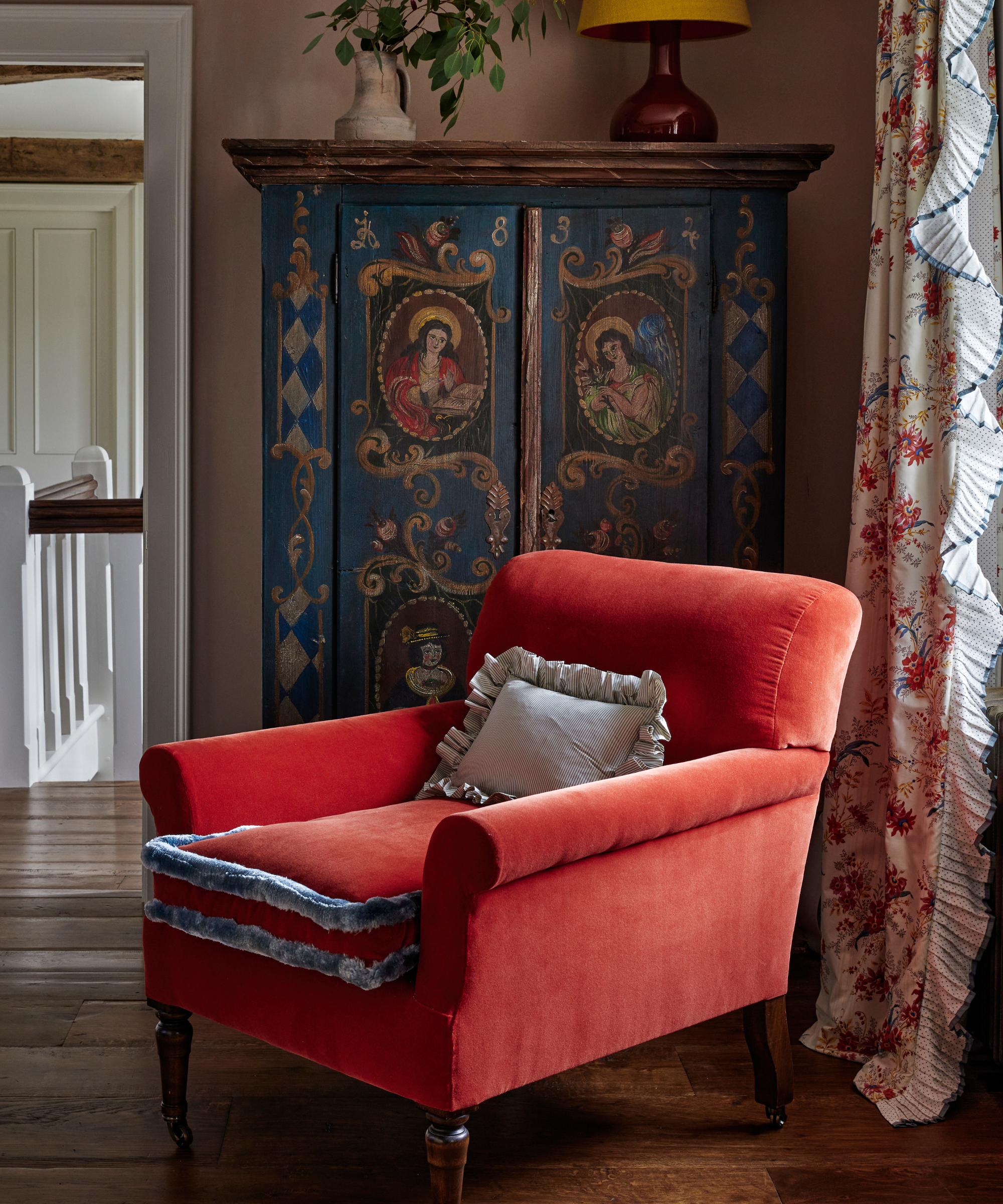 a red accent chair in a room with a vintage painted hutch, a frilly curtain in front of a window, and hardwood floors