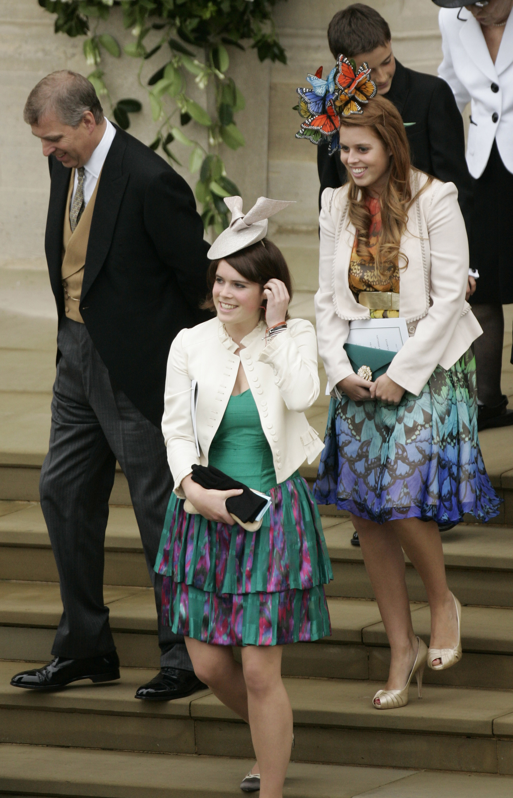 Princess Beatrice and Princess Eugenie wearing bright green and blue dresses with ivory jackets walking down stairs next to Prince Andrew