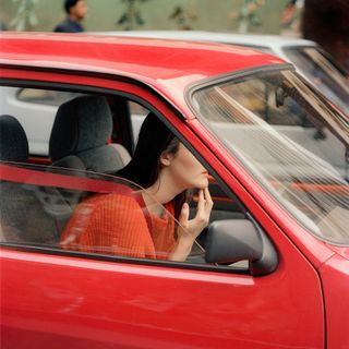 A woman with black hair wearing an orange jersey driving a red car.
