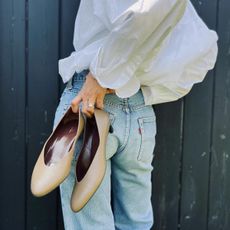 Woman holding up nude court shoes while wearing white button-down shirt and light-wash jeans.