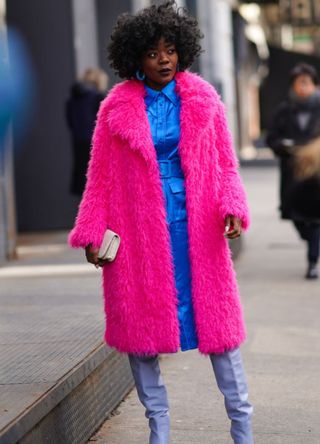 A woman in New York wearing a fuchsia furry coat with a blue dress and lilac boots.