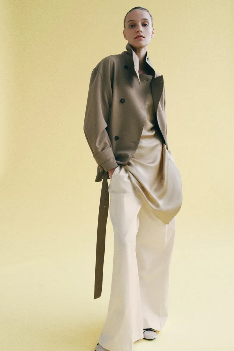Woman wears beige cropped trench coat, silk flowing top and cream trousers